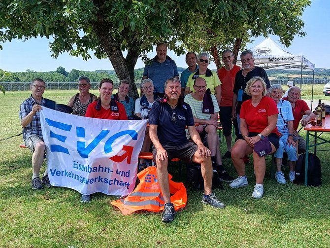 Gruppe von etwa 14 Personen sitzt und steht im Freien unter einem großen Baum auf einer Wiese. Im Vordergrund sitzt eine Person auf einer Bank, daneben halten zwei Personen ein weißes Banner mit blau-rotem Logo und der Aufschrift „EVG Eisenbahn- und Verkehrsgewerkschaft“. Mehrere Personen tragen rote oder dunkle T‑Shirts mit „EVG“-Aufdruck. Auf dem Boden liegt eine orangefarbene Warnweste. Im Hintergrund sind ein Sonnenschirm mit „24MX“-Aufdruck, ein Parkplatz mit Fahrzeugen, Felder und Bäume zu sehen.
