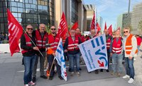 Mehrere Personen stehen auf einem Platz vor modernen Bürogebäuden und halten rote sowie weiße Gewerkschaftsfahnen. Viele Personen tragen rote Warnwesten mit reflektierenden Streifen, eine Person trägt eine orange Warnweste. Im Vordergrund ist eine große weiße Fahne mit blau-rotem EVG-Logo und dem sichtbaren Text „Eisenbahn- und Verkehrsgewerkschaft“. Auf den roten Fahnen sind weiße Schriftzüge und Logos zu sehen. Im Hintergrund befinden sich weitere Personen, Fahnen, Glasfassaden und Hochhäuser bei Tageslicht.