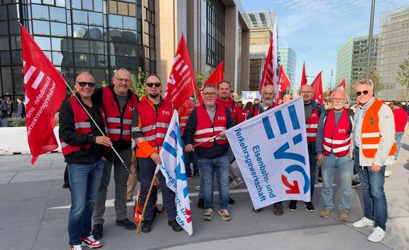 Mehrere Personen stehen auf einem Platz vor modernen Bürogebäuden und halten rote sowie weiße Gewerkschaftsfahnen. Viele Personen tragen rote Warnwesten mit reflektierenden Streifen, eine Person trägt eine orange Warnweste. Im Vordergrund ist eine große weiße Fahne mit blau-rotem EVG-Logo und dem sichtbaren Text „Eisenbahn- und Verkehrsgewerkschaft“. Auf den roten Fahnen sind weiße Schriftzüge und Logos zu sehen. Im Hintergrund befinden sich weitere Personen, Fahnen, Glasfassaden und Hochhäuser bei Tageslicht.