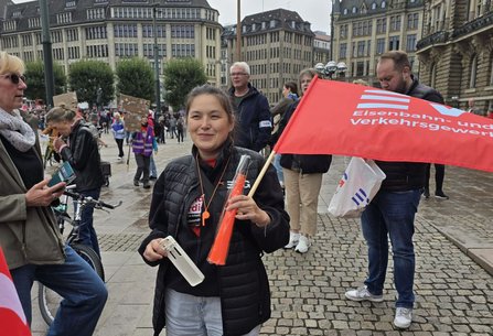 Eine Person steht auf einem gepflasterten Platz und hält eine rote Fahne mit der Aufschrift „Eisenbahn- und Verkehrsgewerkschaft“. In der Hand befinden sich außerdem ein weißes Gerät und ein orangefarbener Gegenstand. Im Hintergrund sind zahlreiche weitere Personen, Fahnen und große historische Gebäude zu sehen.