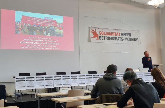 Konferenzraum mit Reihen von Tischen und sitzenden Personen. An der linken Wand ist eine projizierte Präsentationsfolie mit dem Titel „13. Bundeskonferenz ‚Betriebsräte im Visier‘ Gerechtigkeit erkämpfen!“, darunter Datum, Uhrzeit und Ort „Samstag, 10.10.26, 13 Uhr, Gewerkschaftshaus Mannheim“. Rechts hängt ein Banner mit der Aufschrift „SOLIDARITÄT GEGEN BETRIEBSRATS-MOBBING“. Vorne steht eine Person mit Mikrofon neben einem Tisch mit Laptop. Entlang der Vorderseite der Bühne sind mehrere Plakate aufgestellt. Heller Raum mit weißer Wand und Deckenleuchte.