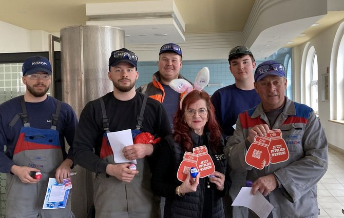 Sechs Personen stehen nebeneinander in einem hellen Innenraum mit Säulen, Fenstern und einer hohen Decke. Mehrere tragen Arbeitskleidung oder Latzhosen in Grau, Blau oder Schwarz mit aufgedruckten Firmenlogos sowie Kappen. In den Händen halten sie rote, schildförmige Karten und Flyer mit der Aufschrift „EVG – JETZT MITGLIED WERDEN!“.