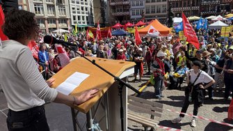  Person steht an einem Rednerpult mit Mikrofon auf einer erhöhten Bühne. Auf dem Pult liegt ein Blatt Papier. Vor der Bühne befindet sich eine große Menschenmenge auf einem Platz mit historischen Gebäuden. Im Hintergrund sind Fahnen, Banner, Zelte und Schirme in verschiedenen Farben zu sehen.