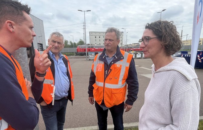 Vier Personen stehen auf einem befestigten Gelände im Freien und sind einander zugewandt. Drei Personen tragen orange Warnwesten mit „DB“-Logo über Alltagskleidung. Im Hintergrund sind Bahngleise, eine rote Lokomotive, Laternenmasten und mehrstöckige Gebäude zu sehen. Der Himmel ist bewölkt.