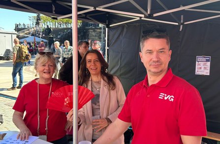 Unter einem schwarzen Pavillon stehen drei Personen an einem Tisch. Zwei Personen tragen rote Oberteile mit weißem „EVG“-Logo, eine Person hält einen roten Fächer. Auf dem Tisch liegen Papiere und Becher. Im Hintergrund sind weitere Personen, Pavillons und eine steinerne Treppenanlage im Freien zu sehen. Rechts am Pavillon hängt ein Plakat mit Text und Prozentangaben.