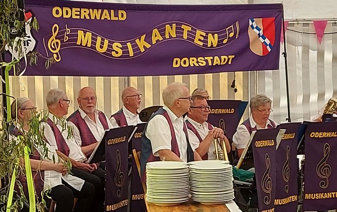 Musikgruppe in einem Festzelt vor einem lila Banner mit der Aufschrift „ODERWALD MUSIKANTEN DORSTADT“. Mehrere Personen in weißen Hemden und dunklen Westen sitzen hinter Notenständern mit der Aufschrift „ODERWALD“. Im Vordergrund steht ein Stapel weißer Teller auf einem Tisch.