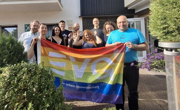 Zehn Personen stehen im Freien vor einem Gebäude und halten gemeinsam eine große Regenbogenflagge mit der Aufschrift „EVG“ und dem teilweise sichtbaren Text „… leben Gemeinschaft“. Im Vordergrund befinden sich Büsche und Blumen, im Hintergrund sind die Gebäudefassade, eine geöffnete Tür und ein grünes Hinweisschild zu sehen. Auf dem blauen T‑Shirt einer Person rechts ist der Text „25. BMT 06.–09.06.2025“ erkennbar.