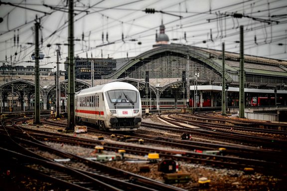 Weißer Zug mit rotem Streifen und sichtbarem „DB“-Logo auf einem Gleis vor einer großen Bahnhofshalle mit bogenförmigem Dach. Im Vordergrund verlaufen zahlreiche Schienen und Weichen, darüber ein dichtes Netz aus Oberleitungen und Masten. Weitere Züge und Bahnsteige sind im Hintergrund zu sehen.