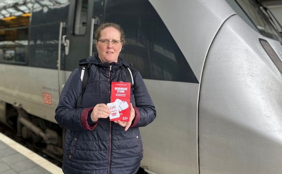 Eine einzelne Person steht auf einem Bahnsteig vor einem silbergrauen Zug. Die Person trägt eine dunkle, gesteppte Jacke und einen Rucksack. In beiden Händen hält die Person ein rotes Faltblatt mit der gut lesbaren Aufschrift „GEMEINSAM STARK“ sowie einem weißen EVG‑Logo. Der Zug zeigt seitlich Fenster, Türen und ein rotes „DB“-Logo. Der Bahnsteig ist gepflastert, die Szene wirkt überdacht durch die Bahnhofskonstruktion.