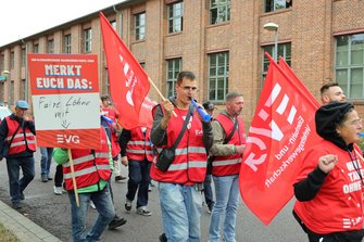 Demonstrationszug auf einer Straße vor einem langgestreckten Backsteingebäude. Teilnehmende tragen rote Warnwesten und rote EVG-Fahnen. Im Vordergrund wird ein Schild mit der Aufschrift „MERKT EUCH DAS: Faire Löhne mit“ getragen. Eine Person hält eine blaue Tröte.