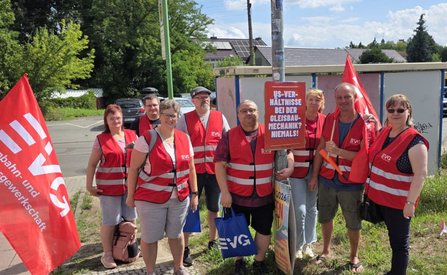 Sieben Personen in roten Warnwesten stehen an einer Straße neben roten EVG-Fahnen. Eine Person hält ein Schild mit der Aufschrift „VS-VERHÄLTNISSE BEI DER GLEISBAUMECHANIK? NIEMALS!“. Mehrere Personen tragen blaue Taschen mit EVG-Logo. Im Hintergrund sind Bäume, geparkte Fahrzeuge und Gebäude zu sehen.