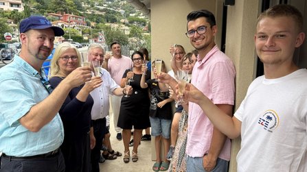 Mehrere Personen stehen vor einem Gebäude und halten Sektgläser in die Höhe. Im Hintergrund sind weitere Personen, geparkte Fahrzeuge, Häuser am Hang und Straßenverkehrsschilder zu sehen. Die Gruppe befindet sich auf einem gepflasterten Außenbereich neben dem Gebäudeeingang.