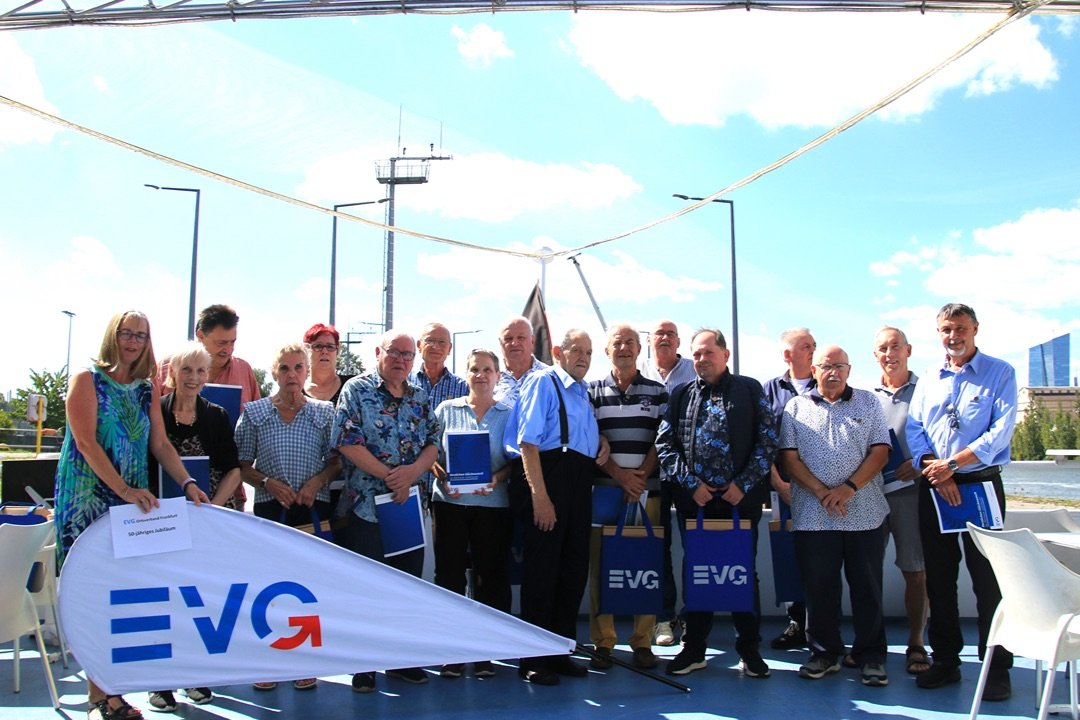 Eine größere Gruppe von Personen steht auf dem Außendeck eines Schiffes hinter einer EVG-Fahne und posiert für ein Gruppenfoto. Die Gruppe umfasst etwa fünfzehn Personen, die dicht nebeneinander auf einem hellblauen Schiffsdeck stehen. Mehrere Personen halten blaue Mappen oder blaue Tragetaschen mit EVG-Logo. Im Vordergrund liegt eine große weiße Fahne mit dem blau-roten EVG-Schriftzug. An der Fahne ist ein Schild befestigt, auf dem „EVG Ortsverband Frankfurt“ und „50-jähriges Jubiläum“ zu lesen ist.