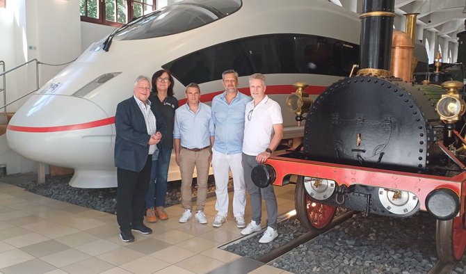Fünf Personen stehen in einer Ausstellungshalle zwischen der Front eines weißen Hochgeschwindigkeitszugs mit rotem Streifen und einer schwarzen historischen Dampflokomotive mit roten Rädern. Der Boden ist gefliest, im Hintergrund sind Fenster und weitere Ausstellungsbereiche sichtbar.