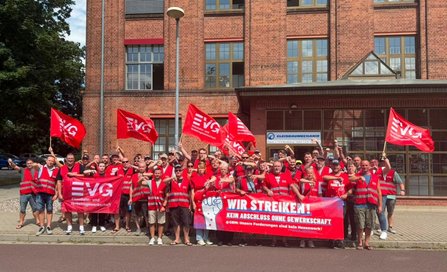 Große Gruppe von Personen vor einem mehrstöckigen Backsteingebäude. Viele tragen rote Warnwesten und halten rote Fahnen mit der Aufschrift „EVG Eisenbahn- und Verkehrsgewerkschaft“. Vor der Gruppe wird ein großes Banner gehalten mit dem Text „WIR STREIKEN! KEIN ABSCHLUSS OHNE GEWERKSCHAFT“. Über einem Eingang ist ein Schild mit der Aufschrift „GLEISBAUMECHANIK“ sichtbar.