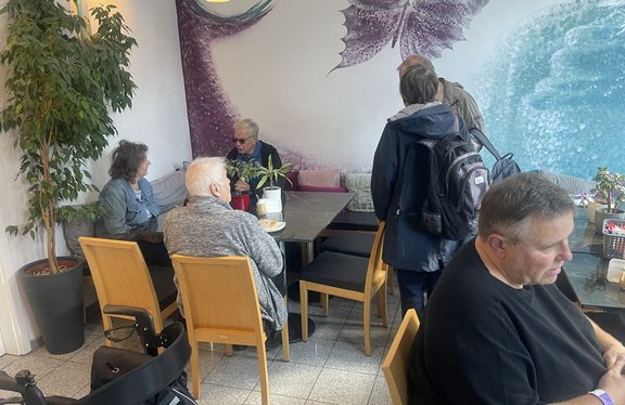 Mehrere Personen befinden sich in einem Innenraum mit Tischen und Holzstühlen. Zwei Personen sitzen an einem Tisch im Hintergrund, eine weitere Person sitzt mit dem Rücken zur Kamera im Vordergrund. Drei Personen stehen rechts neben dem Tisch, eine davon trägt einen Rucksack. Links steht eine große Topfpflanze. Auf den Tischen stehen mehrere kleine Pflanzen, Tassen und Geschirr. Die Wand im Hintergrund ist mit einem großflächigen Wandbild in Violett- und Blautönen gestaltet. Eine Deckenleuchte ist am oberen Bildrand sichtbar.