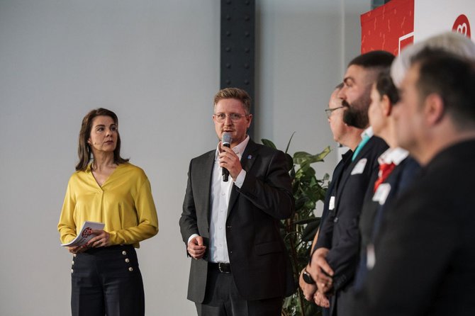 Nahaufnahme eines Bühnenbereichs mit mehreren nebeneinander stehenden Personen. Eine Person hält ein Mikrofon, eine andere ein Kärtchen oder Programmheft. Im Hintergrund sind Pflanzen und Teile der Bühnenkonstruktion sichtbar. Der Hintergrund ist hell.