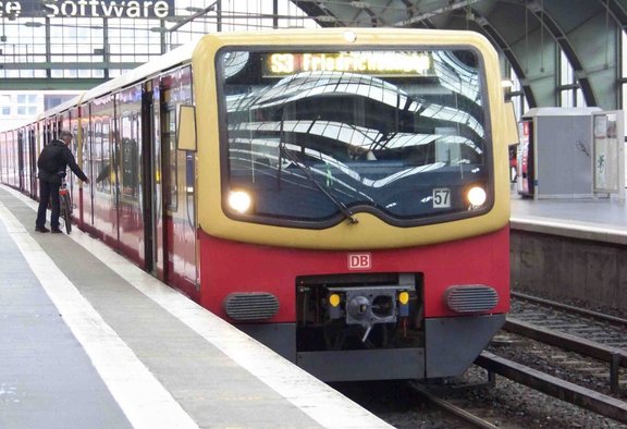 Rote und gelb lackierte S-Bahn mit DB-Logo steht an einem Bahnsteig in einer großen, überdachten Bahnhofshalle mit Glasdach. Die Frontanzeige zeigt „S3 Friedrichst…“. Zwei eingeschaltete Frontlichter sind sichtbar, vorne befindet sich eine Kupplung. Seitlich sind mehrere Türen geöffnet; an einer Tür schiebt eine Person ein Fahrrad in den Zug. Auf dem Bahnsteig sind Bahnsteigkante, Gleise und technische Anlagen zu sehen.