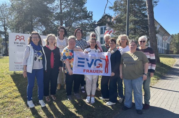 Gruppe von etwa dreizehn Personen, die im Freien auf einer Rasenfläche stehen. In der Mitte halten mehrere Personen ein weißes Banner mit der Aufschrift „EVG FRAUEN“ in blauen und roten Buchstaben sowie einer roten stilisierten Figur. Die Personen tragen unterschiedliche Freizeitkleidung wie T‑Shirts, Pullover, Jacken, Jeans und Stoffhosen in verschiedenen Farben und Mustern. Im Hintergrund sind hohe Bäume, ein helles Gebäude mit Fenstern und Fahnen sowie ein Schild mit der Aufschrift „BSW‑Hotel Ahlbeck“ zu sehen. Rechts verläuft ein gepflasterter Weg mit einem runden Verkehrsschild. Die Szene ist bei sonnigem Tageslicht aufgenommen.