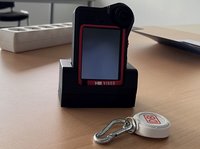 Kleines elektronisches Gerät mit Bildschirm und rotem Rahmen in einer schwarzen Halterung auf einem Tisch; hinten links eine weiße Steckdosenleiste, vorne rechts ein Schlüsselanhänger mit Karabiner und rundem Chip.
