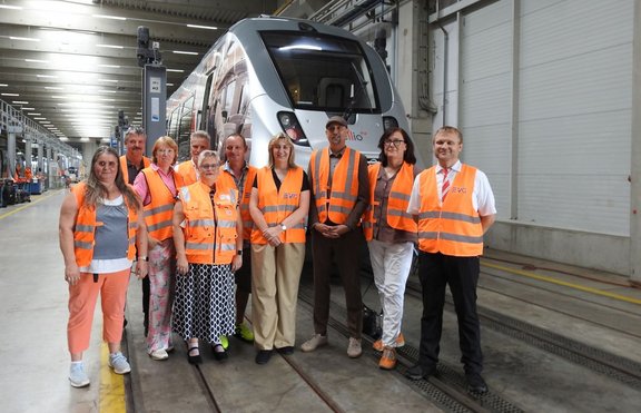 Gruppenfoto in einer Werkhalle vor einem silberfarbenen Schienenfahrzeug. Neun Personen stehen auf den Gleisen beziehungsweise dem Hallenboden und tragen überwiegend orange Warnwesten, teilweise mit sichtbarem EVG-Logo. Im Hintergrund sind die Fahrzeugfront, Wartungsgleise, Hallenstützen, Beleuchtung und weitere Arbeitsbereiche der Halle zu sehen. Die Aufnahme ist gleichmäßig ausgeleuchtet und zeigt die Gruppe frontal vor dem Zug.