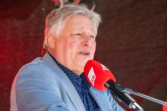 Eine Person steht an einem Rednerpult mit einem Mikrofon mit rotem Windschutz, auf dem „DGB“ steht. Die Person trägt ein hellblaues Sakko über einem dunkelblauen Hemd mit kleinen Punkten. Im Hintergrund sind unscharfe rote Lichtquellen und eine dunkle Fläche zu sehen.