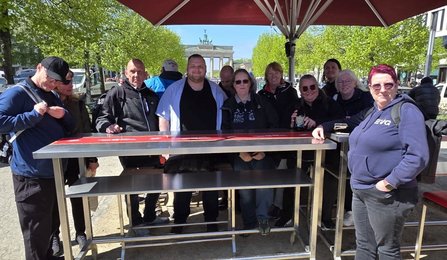 Gruppe von Personen im Freien an einem hohen Metalltisch. Im Hintergrund ist das Brandenburger Tor zu sehen, eingerahmt von Bäumen. Auf dem Tisch liegen Gegenstände; eine Person hält eine Flasche. Ein Sonnenschirm befindet sich über dem Tisch.