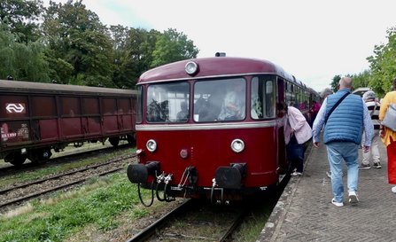 Roter historischer Triebwagen an einem Bahnsteig. Mehrere Personen steigen ein oder gehen entlang des Zuges. Links im Bild stehen offene Güterwagen auf einem parallelen Gleis. Im Hintergrund sind Bäume und weitere Gleisanlagen zu sehen.