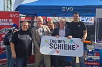 Mehrere Personen stehen vor einem EVG-Informationsstand und halten gemeinsam ein Schild mit der Aufschrift „Tag der Schiene“. Die Gruppe hat sich vor einem blauen Pavillon mit EVG-Logo für ein Foto aufgestellt. Im Mittelpunkt halten mehrere Personen ein großes Schild mit dem Schriftzug „TAG DER SCHIENE“ und einem farbigen grafischen Symbol. Weitere Personen stehen daneben und dahinter.