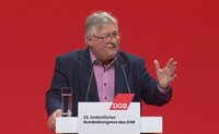 Person hinter einem Rednerpult vor einfarbig rotem Hintergrund. Die Person trägt ein dunkles Jackett und ein helles Hemd. Vor der Person stehen zwei Mikrofone, links auf dem Pult ein Glas mit Wasser. Eine Hand ist seitlich erhoben, die andere liegt am Pult. Auf dem Rednerpult befindet sich ein weißes Schild mit rotem „DGB“-Logo und dem Text „23. Ordentlicher Bundeskongress des DGB“.