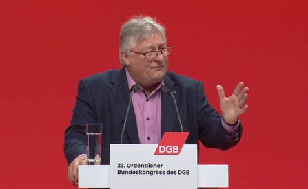 Person hinter einem Rednerpult vor einfarbig rotem Hintergrund. Die Person trägt ein dunkles Jackett und ein helles Hemd. Vor der Person stehen zwei Mikrofone, links auf dem Pult ein Glas mit Wasser. Eine Hand ist seitlich erhoben, die andere liegt am Pult. Auf dem Rednerpult befindet sich ein weißes Schild mit rotem „DGB“-Logo und dem Text „23. Ordentlicher Bundeskongress des DGB“.
