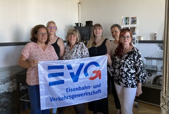 Sechs Personen stehen in einem Innenraum und halten gemeinsam eine weiße Fahne mit blau-rotem EVG-Logo sowie dem Schriftzug „Eisenbahn- und Verkehrsgewerkschaft“. Im Hintergrund sind ein schwarzer Ofen, ein Prospektständer mit Broschüren, große gerahmte Schwarz-Weiß-Fotografien und helle Wände zu sehen.