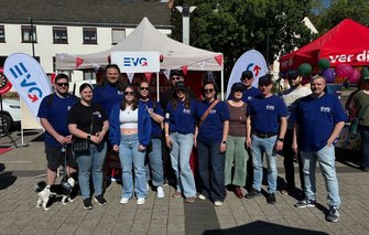 Gruppenfoto im Freien auf einem gepflasterten Platz: Etwa zwölf Personen stehen nebeneinander vor einem weißen Pavillon mit einem Schild „EVG“. Mehrere Personen tragen blaue T‑Shirts mit dem weißen Schriftzug „EVG“. Links und rechts des Pavillons stehen Fahnen mit „EVG“-Logo, im Hintergrund sind weitere Zelte, Ballons, Bäume und ein Gebäude mit Fenstern zu sehen. Im Vordergrund links steht ein kleiner Hund an einer Leine.