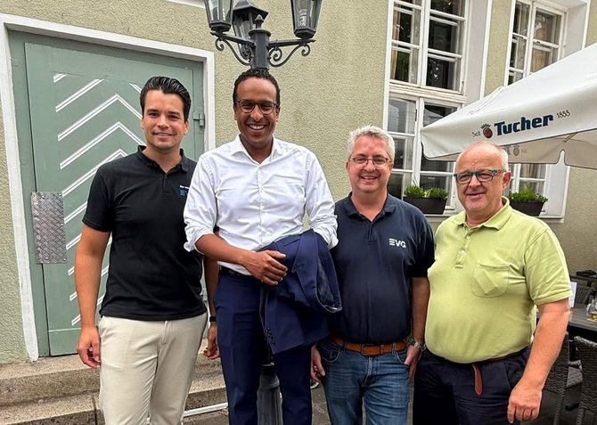 Vier Männer stehen nebeneinander vor einer Hauswand und blicken in die Kamera. Einer trägt ein schwarzes Poloshirt, einer ein weißes Hemd mit Jacke über dem Arm, einer ein dunkles Poloshirt mit Logo und einer ein hellgrünes Poloshirt. Im Hintergrund sind Fenster, eine Tür und ein Sonnenschirm mit „Tucher“-Aufdruck sichtbar.