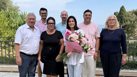 Sieben Personen stehen auf einer Terrasse vor einem Geländer mit Bäumen und Meer im Hintergrund. Eine Person in der Mitte hält einen großen Blumenstrauß mit rosa und weißen Blüten. Die übrigen Personen stehen daneben in einer Reihe. Im Hintergrund sind Vegetation, Himmel und Wasser zu sehen.