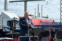 Eine Person spricht auf einer Bühne an einem Bahnhofsgelände in ein Mikrofon, im Hintergrund sind Gleisanlagen und eine Lokomotive zu sehen. Im Vordergrund steht eine Person hinter einem Rednerpult und spricht in ein Mikrofon. Auf der Bühne befinden sich mehrere Mikrofonständer sowie Musikinstrumente, darunter ein Saxofon und ein Kontrabass. Die Person trägt ein Sakko und blickt in Richtung Publikum.