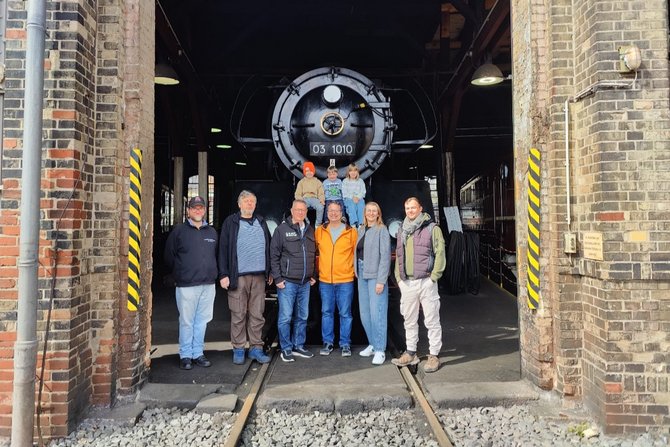 Gruppe von neun Personen (darunter drei Kinder) steht vor einer historischen Dampflokomotive in einer Lokhalle. Die Lok mit der Nummer „03 1010“ befindet sich im Hintergrund auf den Gleisen, eingerahmt von Backsteinmauern und Werkstattelementen. Die Personen blicken in die Kamera, einige stehen auf dem Boden, die Kinder sitzen erhöht vor der Lok.
