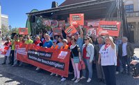 Gruppe von Menschen steht vor einer Bühne auf einem gepflasterten Platz. Mehrere Personen halten rote Schilder mit weißem Text hoch, darunter „Damit die Rente reicht“, „Damit Gesundheit bezahlbar bleibt“ und „Erst unsere Jobs, dann eure Profite“. Im Vordergrund ein breites rotes Banner mit weißem und schwarzem Text: „Hände weg von den Rechten der Beschäftigten!“ und „DGB“, links ein Logo „GEW“ und der Text „Rote Karte dem Kahlschlag!“. Die Bühne hat ein schwarzes Dachgerüst; im Hintergrund Gebäude und Bäume bei sonnigem Tageslicht.