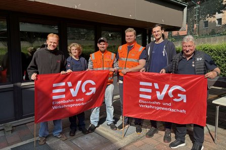 Sechs Personen stehen vor einem Gebäude mit großen Fenstern im Freien. Vier Personen halten zwei rote Fahnen mit der weißen Aufschrift „EVG Eisenbahn- und Verkehrsgewerkschaft“. Zwei Personen tragen orangefarbene Warnwesten. Im Hintergrund sind ein Zaun, Pflanzen und Gebäudeteile zu sehen.