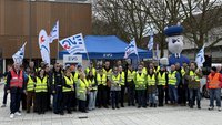 Eine große Gruppe von Menschen steht im Freien vor einem blauen Pavillon. Viele Personen tragen gelbe Warnwesten, einige orangefarbene. Mehrere Personen halten weiß-blaue Fahnen mit EVG‑Logo. Links und rechts sind zusätzliche EVG‑Banner zu sehen. Im Hintergrund steht eine große aufblasbare Figur in blauem Outfit mit Mütze. Hinter der Gruppe befinden sich kahle Bäume und Gebäude.