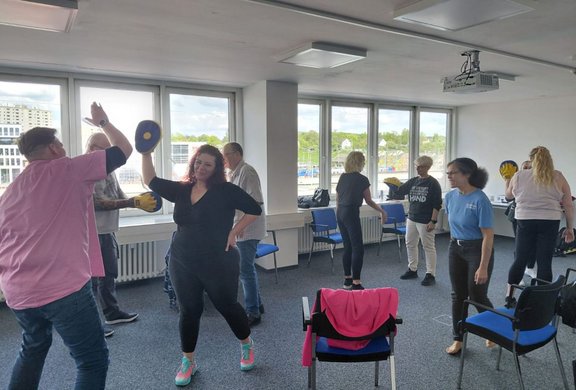 Mehrere Personen befinden sich in einem hellen Seminarraum mit großen Fenstern entlang einer Wand. Einige Personen stehen paarweise gegenüber und halten gelbe Schlagpolster in den Händen, andere führen Bewegungen mit erhobenen Armen aus. Im Raum stehen blaue Stühle, teilweise an den Wänden, ein Stuhl steht mittig im Vordergrund. An der Decke sind Leuchten und ein Deckenbeamer montiert. Durch die Fenster sind Gebäude und ein Außenbereich sichtbar.