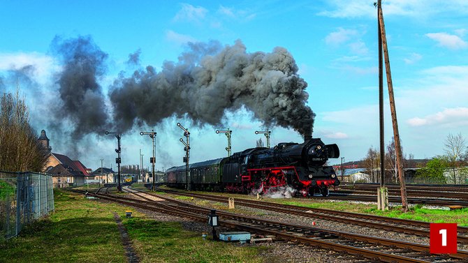 Schwarze Dampflokomotive mit mehreren Personenwagen auf einem Bahngelände mit zahlreichen Gleisen und Signalen. Dichte schwarze und graue Rauchwolken steigen aus dem Schornstein auf. Links stehen Gebäude und Bäume, rechts ein hoher Holzmast. Unten rechts befindet sich eine rote Markierung mit der Zahl „1“.