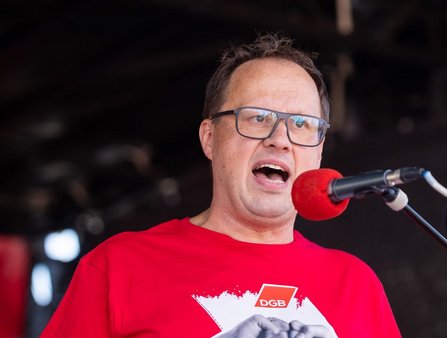 Person mit Brille und rotem T‑Shirt steht vor einem Mikrofon mit rotem Windschutz auf einer Bühne. Auf dem T‑Shirt ist das Logo „DGB“ sichtbar. Der Hintergrund ist dunkel und unscharf.