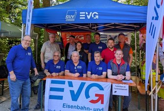 Gruppenfoto im Freien unter einem blauen Pavillon. Auf dem Pavillondach stehen die Logos „GUV FAKULTA“ und „EVG“. Vor dem Pavillon sitzt eine Reihe von Personen an einem Holztisch, weitere Personen stehen dahinter. Mehrere Personen tragen blaue Oberteile mit „EVG“-Aufdruck. Am Tisch hängt ein großes Banner mit der Aufschrift „EVG Eisenbahn- und Verkehrsgewerkschaft“. Rechts am Tisch ist ein Schild angebracht mit dem Text „Toiletten im Turmrestaurant“. Seitlich stehen Fahnen mit EVG-Logo. Im Hintergrund sind Bäume und weitere Zelte zu sehen.