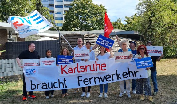 Gruppe von etwa zehn Personen steht im Freien auf einer Wiese vor Bäumen und Wohngebäuden. Die Personen halten Fahnen der EVG sowie mehrere Schilder mit den Aufschriften „Karlsruhe braucht Fernverkehr.“, „Schiene öffnen: Ja. Städte opfern: Nein!“, „Damit Deutschland nicht den Anschluss verliert!“ und „Saarbrücken ohne Fernverkehr? Ohne uns!“. Zwei Fahnen, eine weiß-blaue und eine rote, sind über der Gruppe sichtbar. Sonniges Tageslicht.