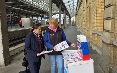 Zwei Personen stehen an einem Bahnsteig neben einem Informationsstand mit Flyern und einem Modell aus großen Kunststoffbausteinen in Rot, Blau und Weiß. Eine Person zeigt auf die ausgelegten Materialien. Im Hintergrund sind Bahngleise, Bahnsteige, Stützpfeiler und das Bahnhofsgebäude zu sehen. Auf dem Modell sind die Wörter „GEMEINSCHAFT“ und „STARKE STIMME“ erkennbar.
