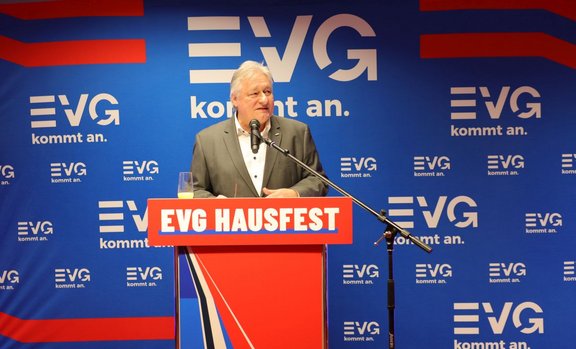Eine Person steht an einem Rednerpult mit der Aufschrift „EVG-HAUSFEST“ und spricht in ein Mikrofon. Hinter dem Rednerpult befindet sich eine blaue Wand mit mehrfach wiederholtem EVG‑Logo und dem Schriftzug „EVG kommt an.“. Auf dem Rednerpult steht ein Glas mit einem Getränk.