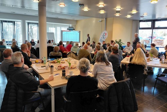Großer heller Veranstaltungsraum mit mehreren rechteckigen Tischen, an denen viele Personen sitzen und nach vorne blicken. Im Vordergrund sind Tische mit Notizblöcken, Stiften, Getränkeflaschen, Kaffeebechern und Tellern mit Gebäck zu sehen. Vorne im Raum steht eine Person neben einer Projektionsfläche, auf der eine Tabelle mit Zahlen und Spalten angezeigt wird, der Text ist nicht lesbar. Rechts neben der Projektionsfläche steht ein Roll‑up‑Banner mit einem roten runden Logo und weißem Schriftzug. Der Raum hat große Fenster an einer Seite, helle Wände, Deckenleuchten und Säulen.