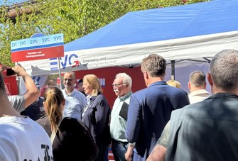 Menschen stehen dicht gedrängt vor einem blauen und weißen Pavillon. Auf dem Pavillon und einem darüber angebrachten Schild ist der Text „EVG Eisenbahn- und Verkehrsgewerkschaft“ zu lesen. Mehrere Personen sind von hinten oder seitlich zu sehen, einige im Gespräch. Links hält eine Person ein Smartphone hoch. Im Hintergrund sind Bäume und industrielle Rohrleitungen sichtbar.