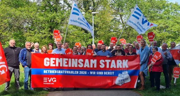 Eine Gruppe von Menschen steht im Freien vor grünen Bäumen und hält ein großes rotes Banner. Auf dem Banner steht in weißer und blauer Schrift: „GEMEINSAM STARK“ und darunter „BETRIEBSRATSWAHLEN 2026 – WIR SIND BEREIT“. Links unten ist das Logo „EVG kommt an.“ zu sehen, rechts ein Symbol mit der Aufschrift „#BRW2026“. Mehrere Personen halten rote Schilder mit der Aufschrift „EVG“ sowie weiße Fahnen mit blauem „EVG“-Logo und dem Text „Eisenbahn- und Verkehrsgewerkschaft“. Die Szene ist bei Tageslicht auf einem Weg oder einer Grünfläche aufgenommen.